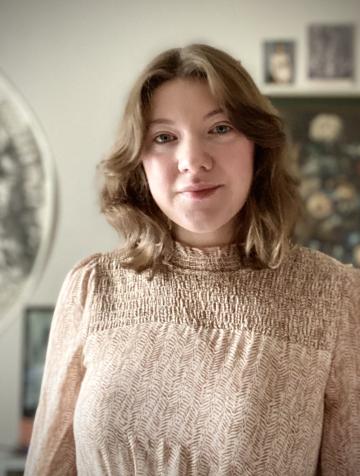 White woman with short dark blonde hair standing before a backdrop with various artworks 