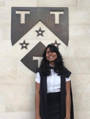 Woman in white shirt, black bowtie and black skirt, smiling and posing beside an Oxford college crest. 