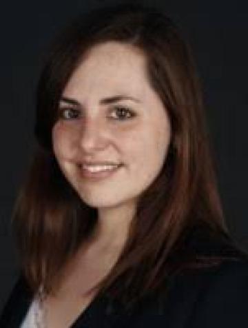 Headshot of white woman with brown hair in black suit jacket with dark background.