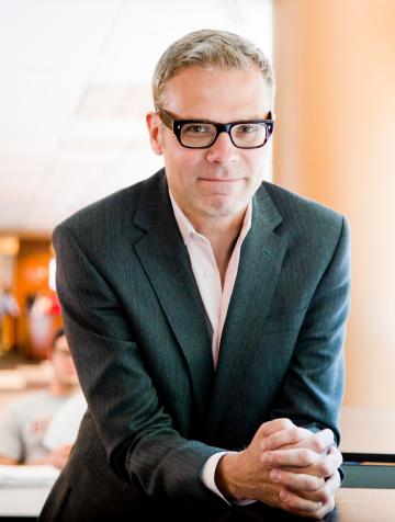 Teemu Ruskola looks into the camera smiling with his hands clasped in front of him. He is wearing glasses and a dark suit.