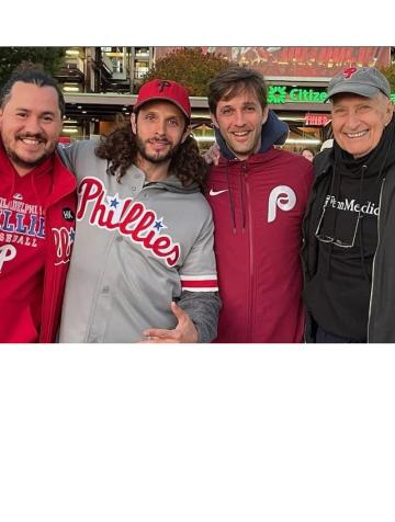 Four people in Philadelphia sports attire stand facing the camera smiling, with arms around each other. To the far right is Bruce Kuklick in a black shirt and gray cap.