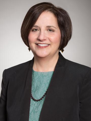 Beth Wenger looks at the camera, smiling. Her hair is dark and chin-length and she is wearing a light blue-green top with a black blazer. 