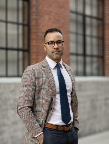 William Sturkey stands in front of a brick building. He is wearing glasses, a tan blazer, and navy tie and pants. 