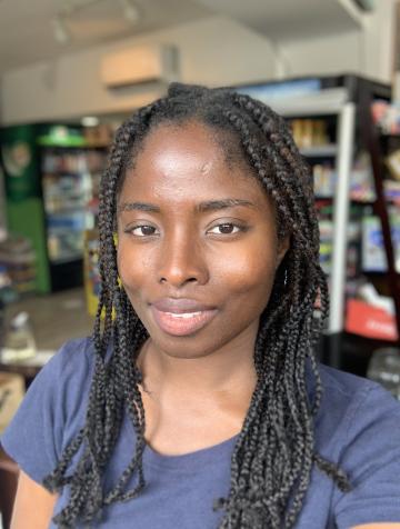dark skinned woman with full lips, high cheekbones, and shoulder length box-braids in a blue t-shirt, with a grocery store behind her