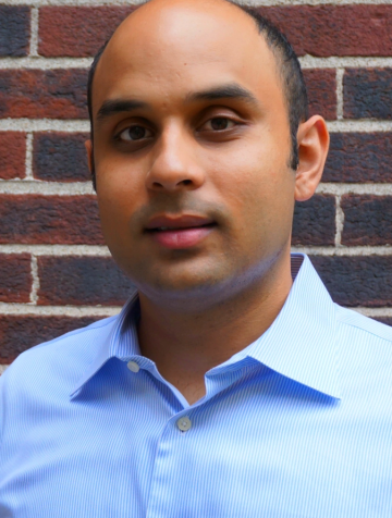 Sudev Sheth is wearing a blue button-down shirt and is standing in front of a brick wall. 