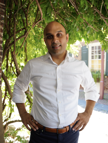 Sudev Sheth is wearing a white button-down shirt in front of hanging greenery.